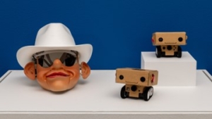 Três pequenas esculturas sobre um pedestal branco. A primeira é uma cabeça de marioneta com um chapéu de cowboy branco e as outras duas são pequenos robots de cartão sorridentes sobre rodas de plástico. Alguns BlabDroids de cartão expostos ao lado de uma máscara de plástico de uma peça de arte performativa, intitulada Five Dollars Can Save Planet Earth.